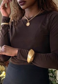 Femme portant un haut à manches longues brun foncé, un collier chaîne en or avec pendentif, et plusieurs bracelets en or, debout à l'intérieur près de plantes et de fleurs.