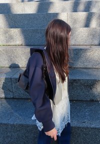 Persona con cabello largo y castaño, vestida con chaqueta azul marino, blusa blanca de encaje y pantalones oscuros, de pie sobre peldaños de piedra gris bajo la luz del sol.