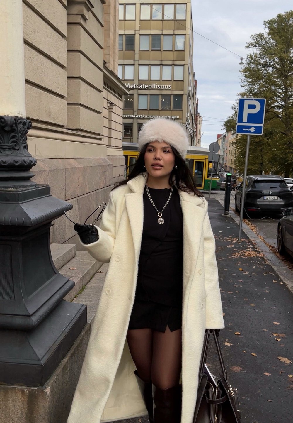 Cappotto in lana bianca dalla texture morbida, abito aderente nero, guanti neri e cappello di pelliccia chiara. Tiene occhiali da sole con una borsa scura al fianco.