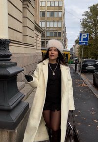 Cappotto in lana bianca dalla texture morbida, abito aderente nero, guanti neri e cappello di pelliccia chiara. Tiene occhiali da sole con una borsa scura al fianco.