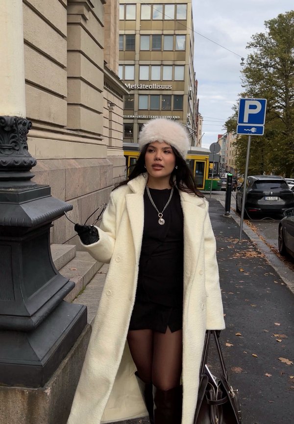 Hvid uldfrakke med en blød tekstur, sort tætsiddende kjole, sorte handsker og en lys pels hat. Holder solbriller med en mørk håndtaske ved siden af sig.