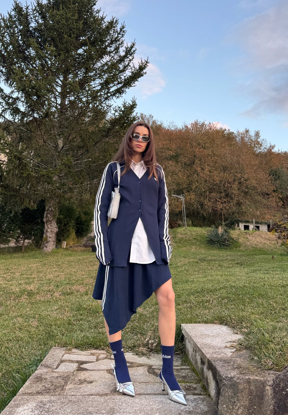 Femme portant une jupe asymétrique bleu marine, une veste à manches longues assortie avec des rayures blanches, une chemise blanche, des talons argentés et des lunettes de soleil à l'extérieur.