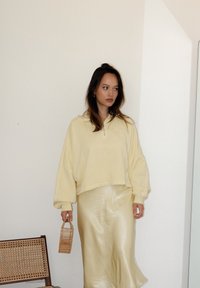 Κίτρινο κοντό φούτερ με υπερμεγέθεις μανίκια, συνδυασμένο με μια γυαλιστερή χρυσή σατέν φούστα. Κρατάει μια μικρή υφασμένη ξύλινη τσάντα.