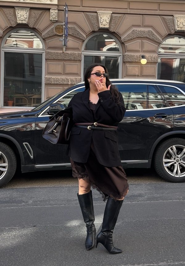 Mujer con blazer negro, falda asimétrica y botas hasta la rodilla, de pie en la calle cerca de un SUV negro, tocándose la cara y llevando gafas de sol.