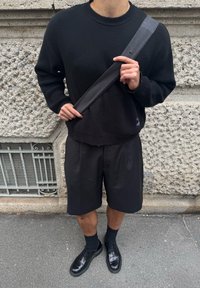 Άνδρας με μαύρο πουλόβερ, μαύρα σορτς μέχρι το γόνατο, μαύρα παπούτσια και μαύρο λουρί τσάντας χιαστί σε αστικό περιβάλλον.