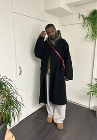 Homme debout à l'intérieur portant un pardessus noir, un sweat à capuche olive, un pantalon de jogging gris, des chaussons marron et un sac bandoulière rouge près de plantes en pot.