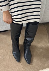 Personne portant des bottes noires à bout pointu et hauteur genou, des collants noirs, et une robe en maille rayée blanc et noir avec des boutons sur la manche.