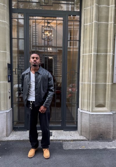 Chaqueta de cuero negra sobre una camisa blanca, combinada con pantalones negros y botas marrones. De pie frente a una puerta con marco metálico decorativo.