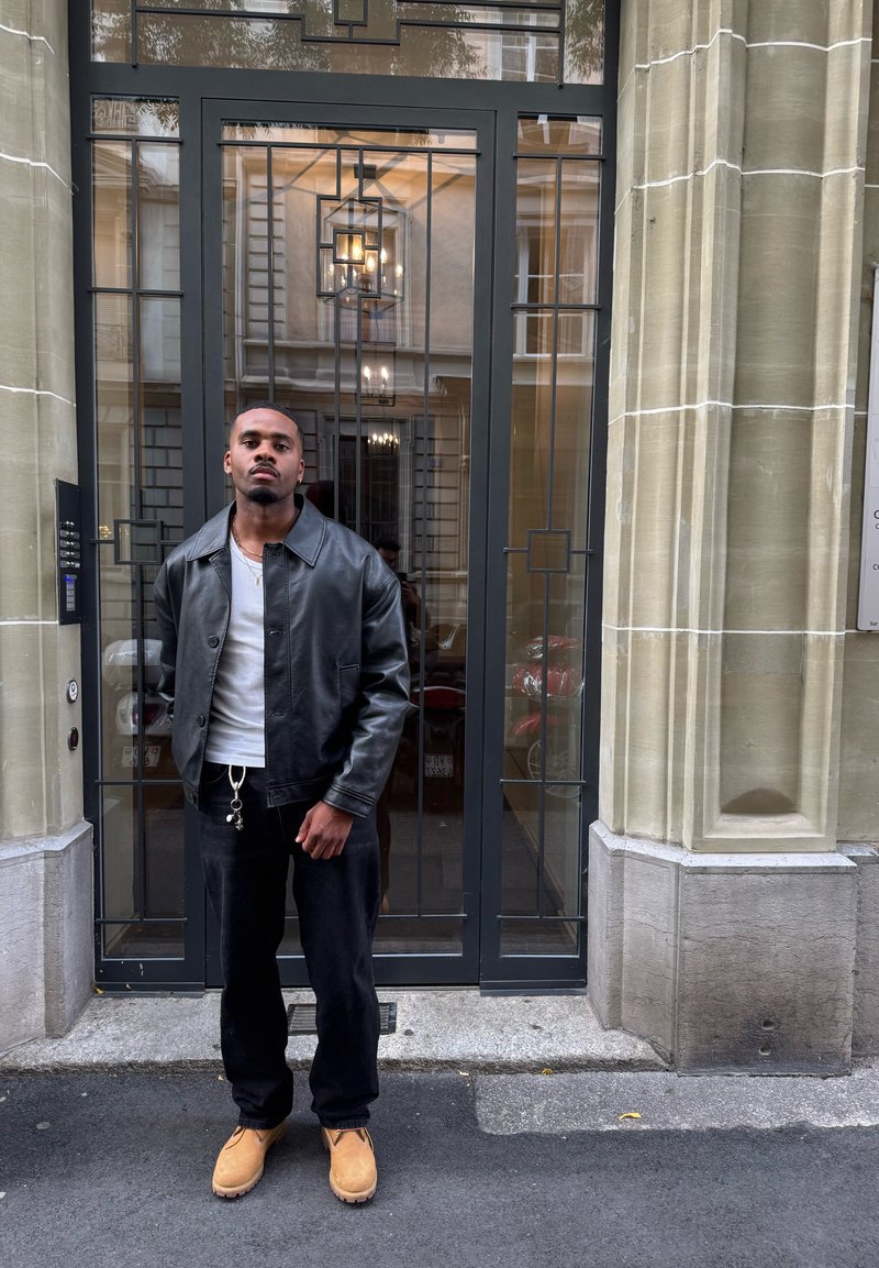 Giacca di pelle nera sopra una camicia bianca, abbinata a pantaloni neri e stivali marroni. In piedi davanti a una porta decorativa con cornice in metallo.