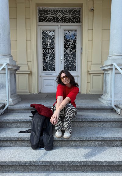 Mujer con top rojo y pantalones con estampado de cebra sentada en escalones de piedra con chaqueta negra y bufanda roja a su lado, delante de puertas dobles blancas ornamentadas.