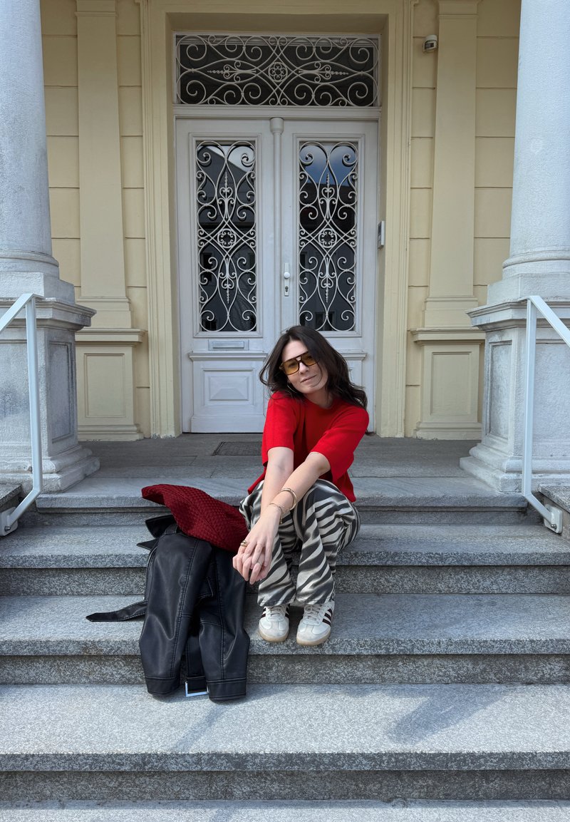 Γυναίκα με κόκκινο τοπ και παντελόνι με ζέβρα, καθισμένη σε πέτρινα σκαλοπάτια με μαύρο μπουφάν και κόκκινο κασκόλ δίπλα της, μπροστά από περίτεχνες λευκές διπλές πόρτες.