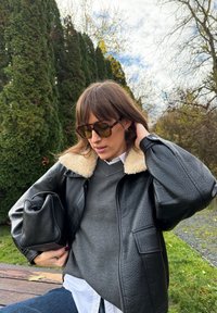 Crna kožna jakna s ovratnikom od ovčje kože, sivi džemper, bijela košulja i tamne sunčane naočale; drži malu crnu torbu. Prirodni vanjski ambijent.