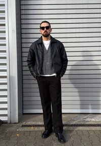 Črna usnjena jakna čez sivi teliran pulover, belo majico s kratkimi rokavi in črne hlače. Oseba stoji ob črtasti kovinski steni.