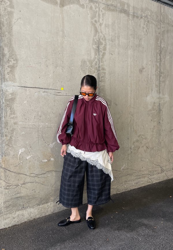 Persona con chaqueta deportiva Adidas color granate, pantalones anchos de cuadros en blanco y negro, zapatos negros sin cordones y gafas de sol naranjas, de pie junto a una pared de hormigón.