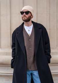 Manteau en laine noir sur un pull marron côtelé, jean clair et bonnet en tricot crème. Accessoires comprenant des colliers superposés et des lunettes de soleil foncées.