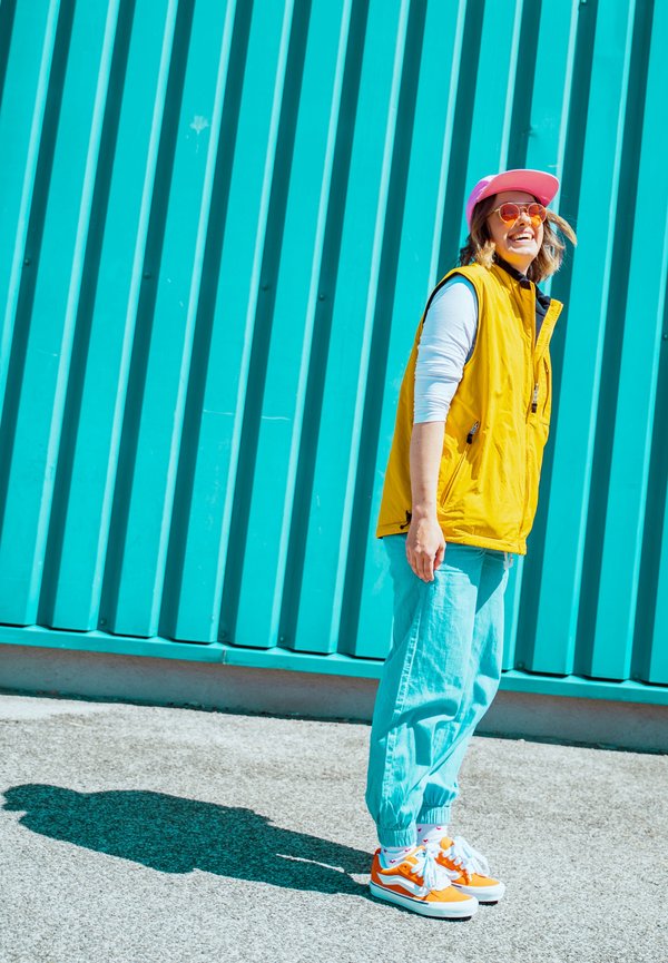 Personne portant un gilet jaune, une casquette rose, des lunettes de soleil orange, un pantalon bleu clair et des baskets orange, debout souriante devant un mur métallique ondulé couleur sarcelle.