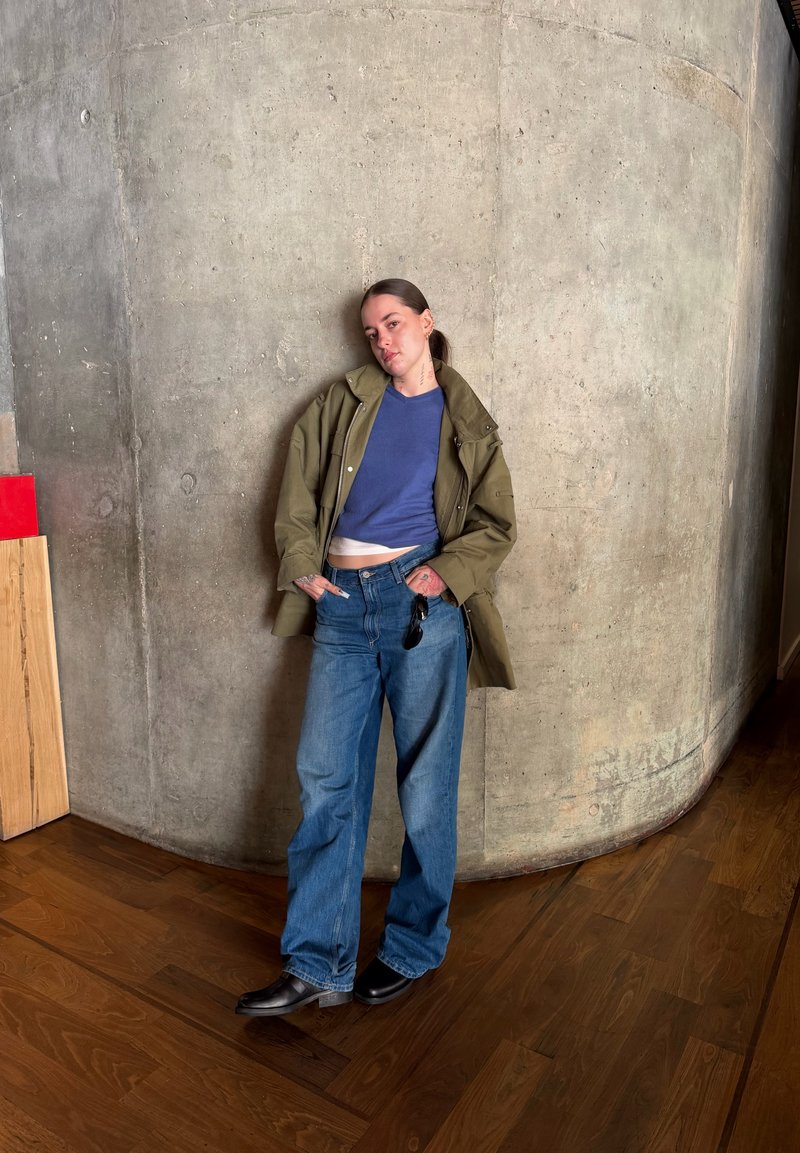 Giovane con tatuaggi, indossa una giacca oliva, una camicia blu, jeans larghi e scarpe nere, appoggiato a una parete curva di cemento all'interno.