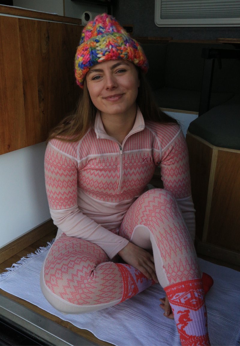 Jeune femme assise en tailleur à l’intérieur, vêtue d’un haut zippé rose à motifs, de leggings assortis, d’un bonnet tricoté coloré et de chaussettes rouges à motifs sur un tapis blanc.