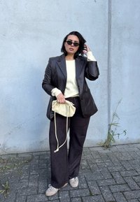 Black leather blazer over a cream sweater, paired with dark pants and light sneakers. Holding a small beige drawstring bag. Concrete background.