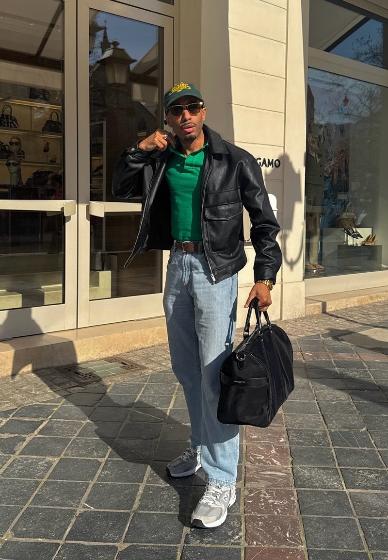 Homme portant une veste en cuir noire, une chemise verte, un jean bleu clair, des lunettes de soleil et une casquette, portant un sac noir devant un commerce dans un cadre urbain ensoleillé.