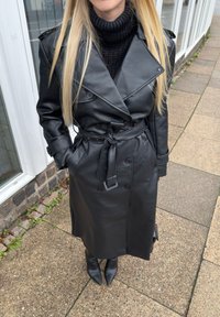 Trench-coat en cuir noir avec une taille ceinturée, des boutons croisés et des poches latérales, accompagné de bottines noires ajustées à talons.
