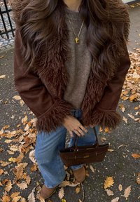 Casaco de camurça castanho com um colarinho fofinho, combinado com um suéter bege, calças de perna larga e uma mala castanha. Acentuado com joias douradas.