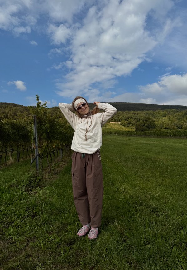 Svetlo bež dolg rokav hoodie, rjave široke hlače, roza obutev z vzorcem. Stoji na travnati površini z brinavkami in modrim nebom.