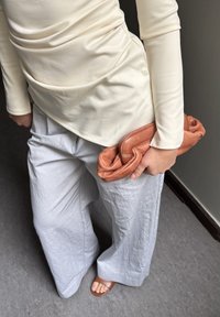 Kreminė ilgomis rankovėmis palaidinė, laisvos pilkos plačios kelnės su tekstūruotu paviršiumi ir oranžinė odinė rankinė laikoma vienoje rankoje.