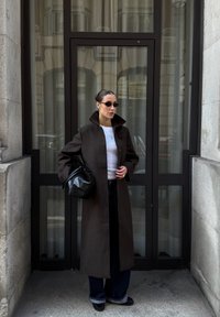 Lange, donkerbruine wollen jas met hoge kraag, gedragen over een witte top en brede, donkere jeans. Geverfde zwarte handtas en zonnebril.