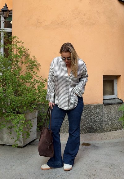 Blusa de cuadros blancos y negros, jeans oscuros de pierna acampanada, bolso tote marrón y zapatillas blancas. Entorno al aire libre con pared color naranja pálido.