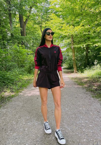 Chándal deportivo negro con detalles en rosa, chaqueta con cremallera y pantalones cortos. Combinado con zapatillas negras y blancas, ambientado en un área boscosa verde.