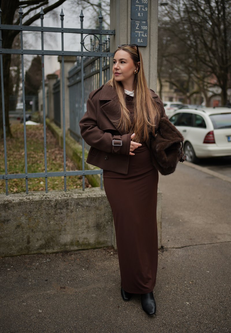 Femeie îmbrăcată într-un costum maro, ținând o haină de blană, stând lângă o gardă metalică pe o stradă urbană, cu mașini parcate în fundal.