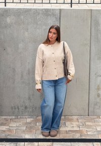 Beige kötött kardigán gombokkal, világoskék, széles szárú farmernadrággal párosítva. Szürke válltáskával és barna cipővel kombinálva. Textúrált háttér.