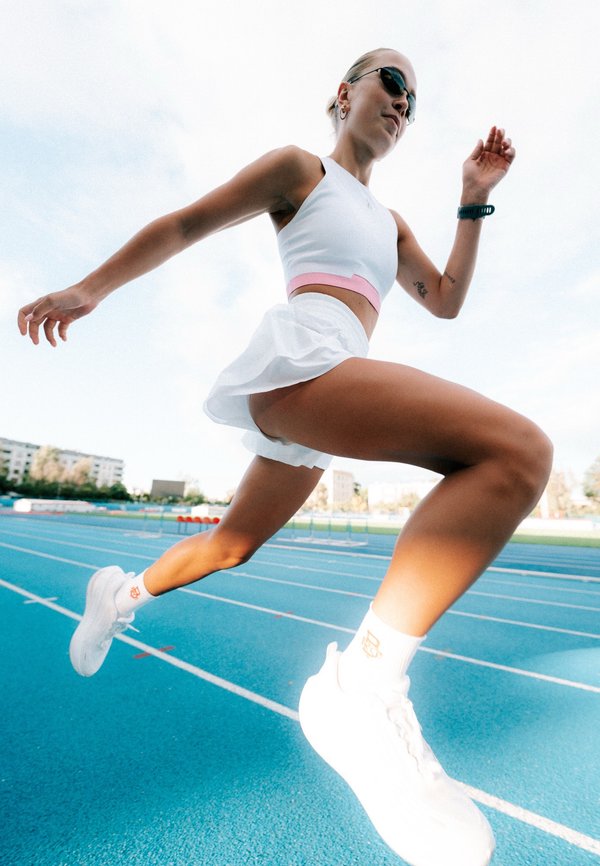 Athlète féminine en tenue de sport blanche et lunettes de soleil courant sur une piste extérieure bleue en plein jour.