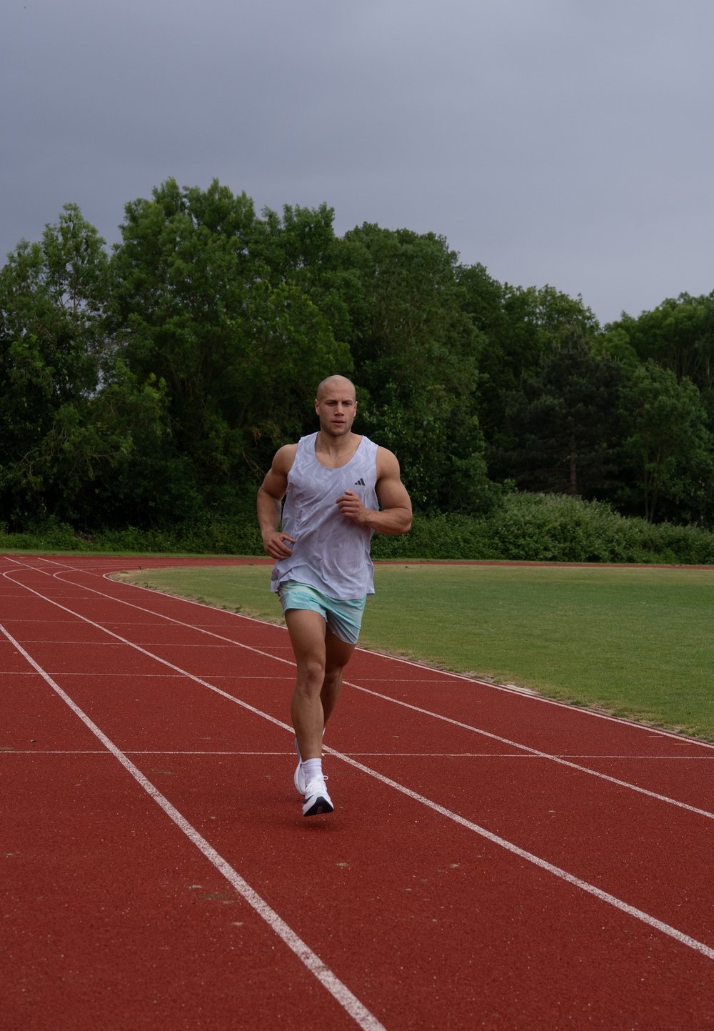 Idrettsutøver som løper på en rød bane, ikledd en ermeløs grå skjorte og lyseblå shorts, med hvite joggesko. Grønne trær i bakgrunnen.