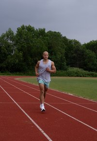 Idrettsutøver som løper på en rød bane, ikledd en ermeløs grå skjorte og lyseblå shorts, med hvite joggesko. Grønne trær i bakgrunnen.