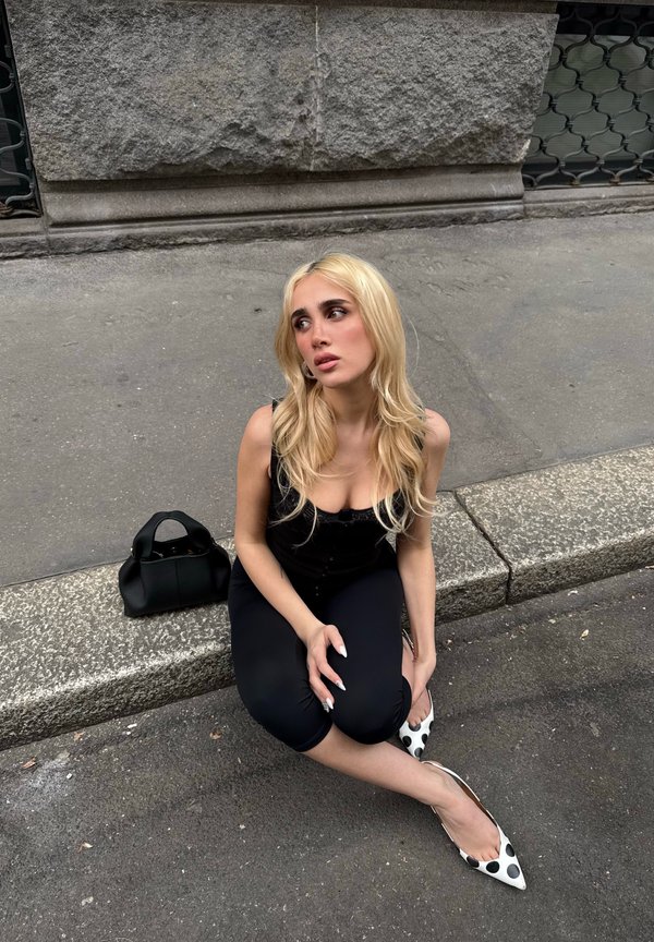 Jeune femme aux longs cheveux blonds vêtue d'une tenue noire et de chaussures à pois, assise sur un rebord en pierre à côté d'un sac à main noir sur le trottoir.