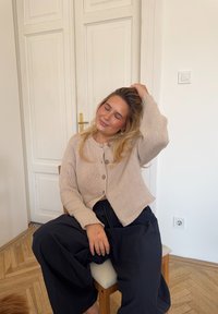 Cardigan bej tricotat, cu guler rotund și nasturi din lemn, purtat cu pantaloni negri largi, așezat pe un scaun pe un fundal deschis.