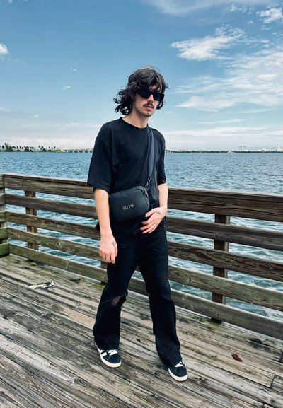 Bolso bandolera negro suave con el logo "arte", llevado sobre una camisa y pantalones negros. Modelo en un muelle de madera con agua y cielo de fondo.