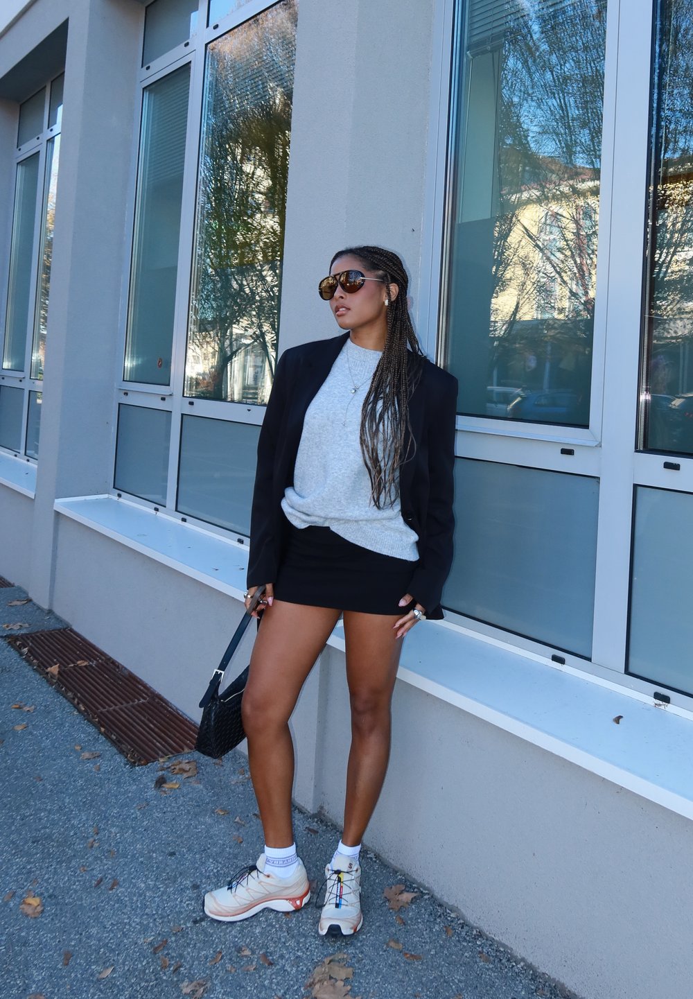 Zwarte blazer, grijze trui, zwarte minirok en beige sneakers. Accessoires zijn onder andere grote zonnebril en een kleine zwarte handtas.