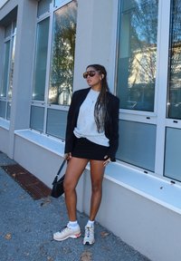 Zwarte blazer, grijze trui, zwarte minirok en beige sneakers. Accessoires zijn onder andere grote zonnebril en een kleine zwarte handtas.