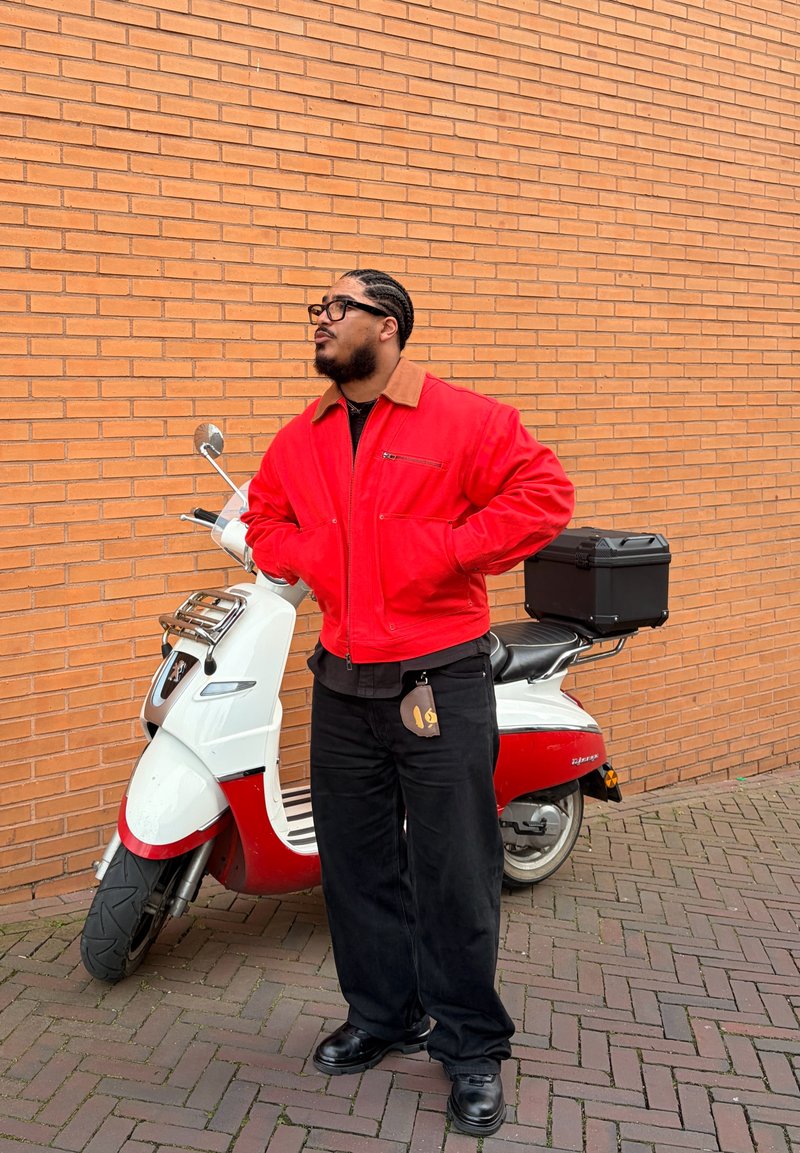 Homme aux cheveux tressés et lunettes portant une veste rouge se tient les mains dans les poches à côté d'un scooter blanc et rouge garé contre un mur de briques.
