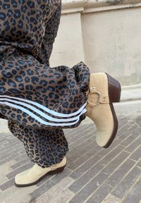 Bottines en daim beige avec talons en bois portées avec un pantalon à imprimé léopard marron avec des bandes blanches sur les côtés, sur une rue pavée.