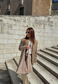 Blazer oversized bege usado sobre uma camisa branca, combinado com calças creme. Bolsa tote grande em camurça macia numa tonalidade correspondente. Degraus de pedra ao fundo.