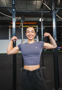Grå atletisk crop-top med korte ermer, vist med bøyd biceps; svarte treningsbukser; treningsstudio med utstyr i bakgrunnen.