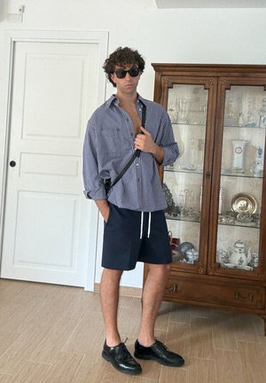 Camisa de gran tamaño a cuadros en azul y blanco, pantalones cortos azul marino, zapatos negros, usando gafas de sol, con un bolso cruzado y un mueble de madera de fondo.