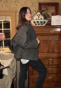 Veste grise oversize à finition texturée, superposée à une chemise blanche. Sac noir porté à l'épaule, debout devant un meuble en bois.