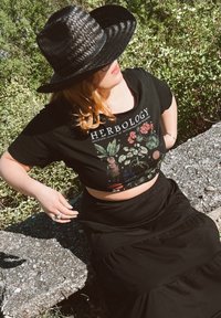 Μαύρο κοντό μπλουζάκι με γραφικό «Φυτολογία», συνδυασμένο με μια μαύρη μακριά, ροή φούστα. Μεγάλο πλεκτό μαύρο καπέλο ηλίου. Υπαίθριο περιβάλλον.