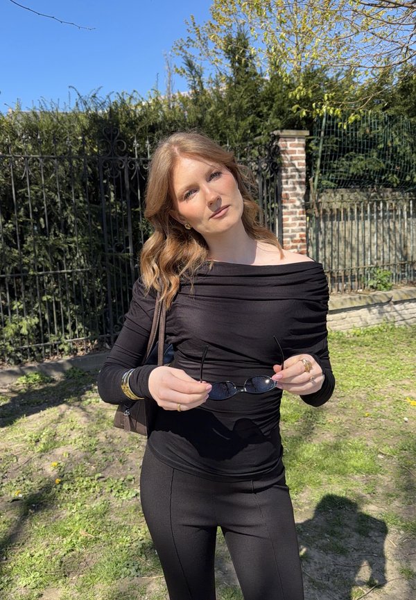 Jonge vrouw met lichtbruin haar in een zwarte off-shoulder top en broek staat buiten en houdt een zonnebril vast, met een in de zon gelegen tuin en een ijzeren hek op de achtergrond.