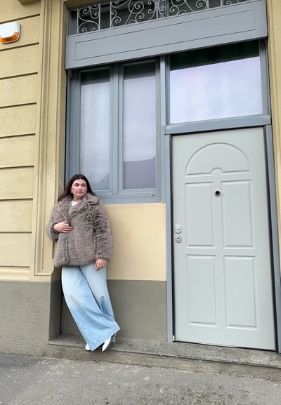 Abrigo de piel sintética en beige, jeans de corte holgado en azul claro, de pie junto a una puerta gris con un pomo redondo, fondo de pared texturizada, grandes ventanales.